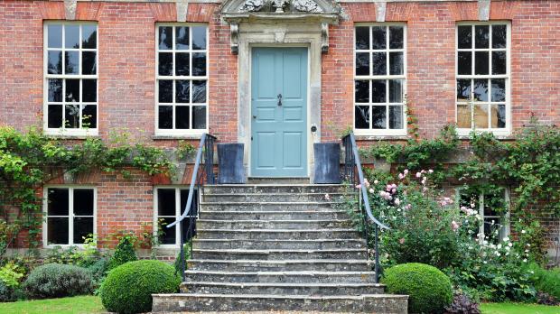The front of a red brick country house, with large windows and a pale blue door at the top of a set of stone steps leading into a manicured garden